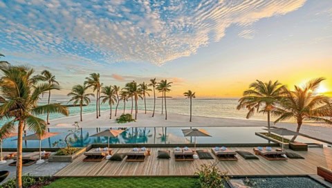 Infinity-Pool mit Sonnenliegen und Blick auf weißen Sandstrand und türkisfarbene Lagune bei Sonnenuntergang