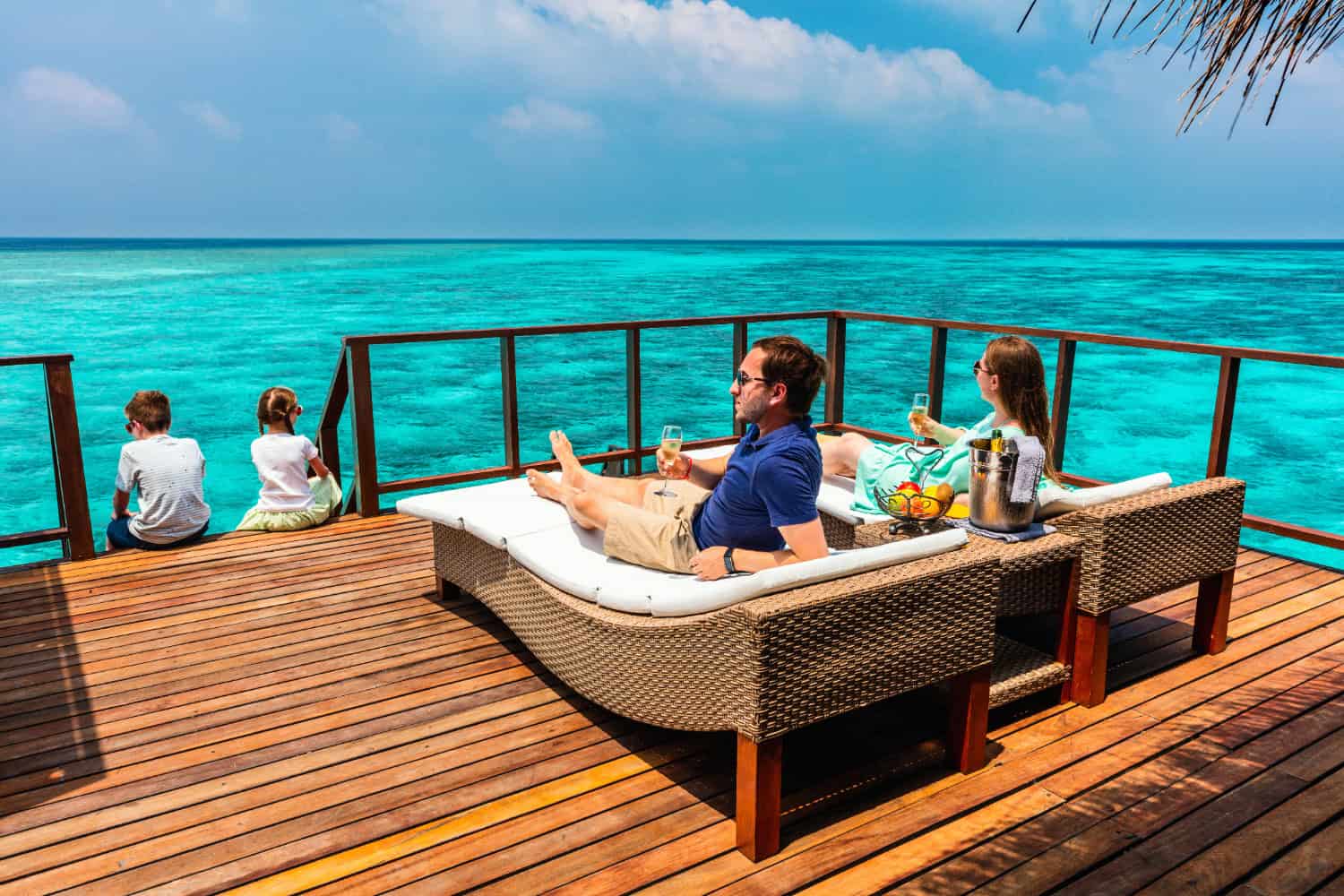 Familie entspannt auf Terrasse eines Wasserbungalows mit Blick auf das türkisblaue Meer