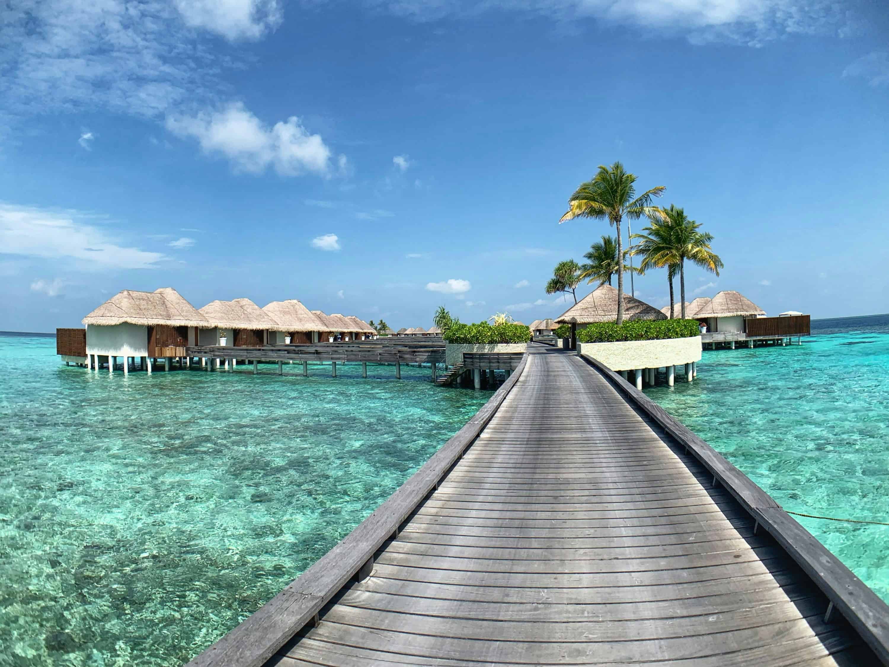 Holzsteg führt über türkisfarbenes Wasser zu Wasserbungalows auf den Malediven unter blauem Himmel