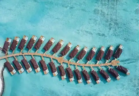 Luftaufnahme von Overwater-Villen in geschwungener Form über türkisblauem Meer.
